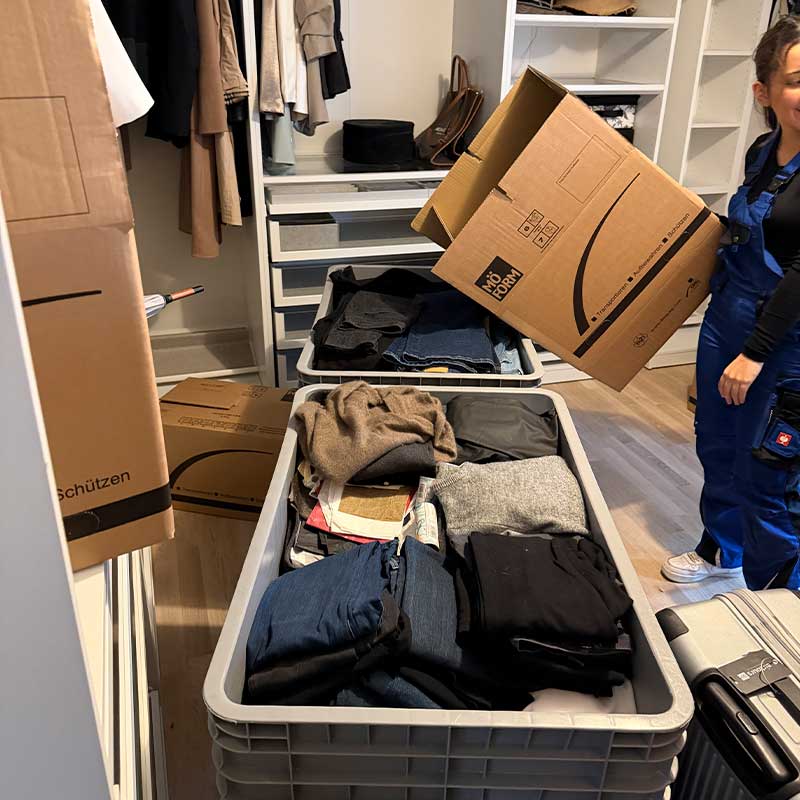 Umzugshelfer verpacken Kleidung und persönliche Gegenstände in Transportboxen innerhalb eines Ankleidebereichs.
