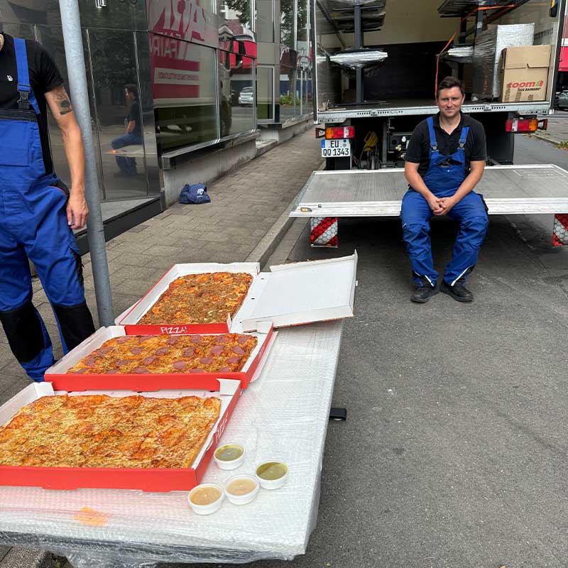 Umzugshelfer machen während eines Umzugs eine Pause und essen Pizza vor dem geöffneten Umzugsfahrzeug.