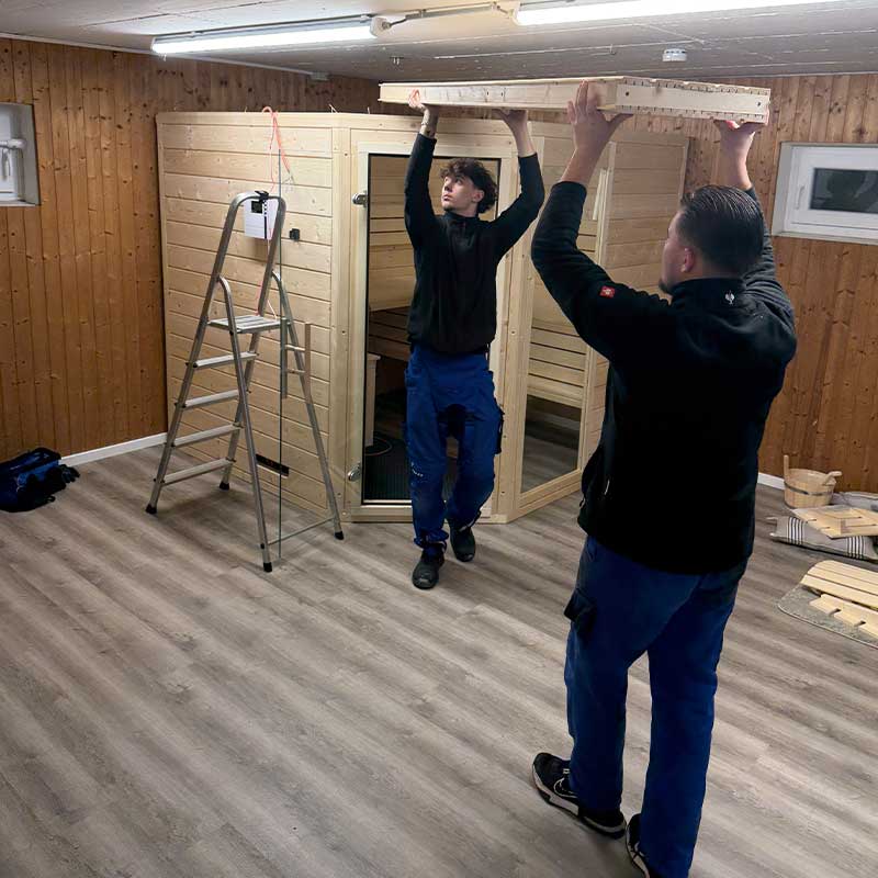 Zwei Monteure montieren eine Holzsauna und setzen das obere Bauelement fachgerecht ein.